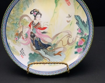 May include: A decorative porcelain plate with a gold trim and a colourful image of a woman in a flowing gown with a fan. The plate features a mountain scene with flowers and butterflies. The plate has Chinese characters on it.