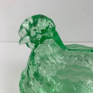 May include: A green glass hen-shaped dish with a textured surface. The hen is depicted in a sitting position, with detailed feather patterns. The dish is likely used for decorative or serving purposes.