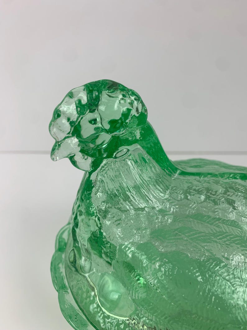 May include: A green glass hen-shaped dish with a textured surface. The hen is depicted in a sitting position, with detailed feather patterns. The dish is likely used for decorative or serving purposes.