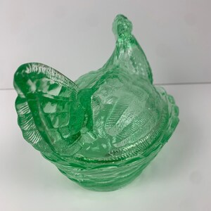 May include: A green glass hen-shaped dish with a textured surface. The dish is a light green color and has a detailed design, including feathers and a ruffled edge. The lid of the dish is shaped like the hen's head.