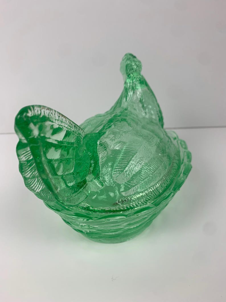 May include: A green glass hen-shaped dish with a textured surface. The dish is a light green color and has a detailed design, including feathers and a ruffled edge. The lid of the dish is shaped like the hen's head.