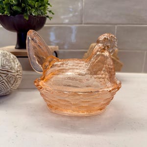 May include: A peach-colored glass hen-shaped dish with a textured surface. The hen sits atop a woven basket-like base. The dish is likely vintage and adds a touch of farmhouse charm to any kitchen or dining room.