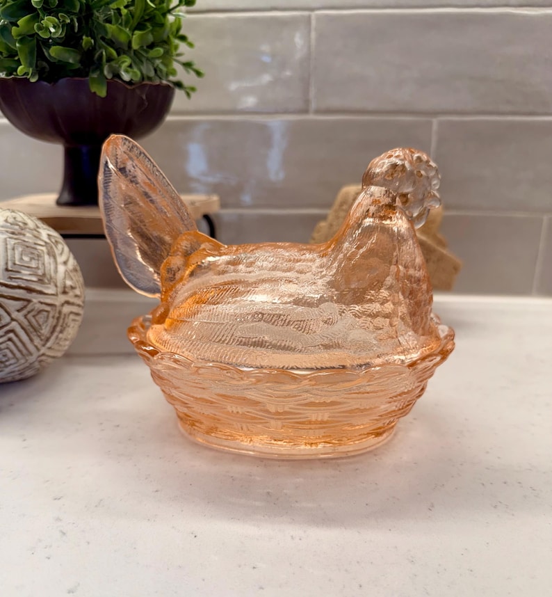 May include: A peach-colored glass hen-shaped dish with a textured surface. The hen sits atop a woven basket-like base. The dish is likely vintage and adds a touch of farmhouse charm to any kitchen or dining room.