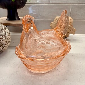 May include: A peach-colored glass hen-shaped dish with a textured surface. The hen sits atop a basket-like base. Decorative item for kitchen or home decor.