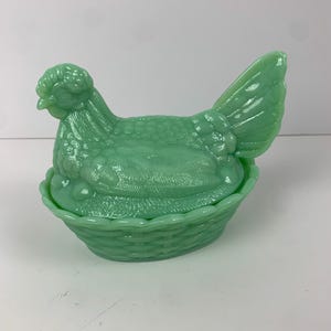May include: A vintage green glass chicken in a nest dish. The chicken is sitting in a woven basket with a lid.