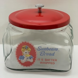 Op de afbeelding: Een vintage glazen broodpot met een rood metalen deksel en een knop. De pot heeft een etiket met de tekst "Sunbeam Bread It's Batter Whipped" en toont een cartoonmeisje dat een snee brood vasthoudt.