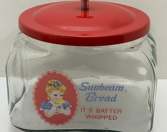 May include: A vintage glass bread jar with a red metal lid and a knob. The jar has a label that says "Sunbeam Bread It's Batter Whipped" and features a cartoon girl holding a slice of bread.