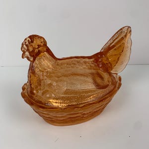 Puede incluir: Un plato vintage de cristal color ámbar con forma de gallina sobre nido. La gallina presenta detalles de plumas texturizadas y una cresta, posada sobre una base de tejido de cesta. Esta pieza decorativa vintage es un clásico de colección.
