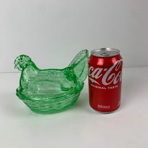 May include: A green glass hen-shaped dish with a textured surface. The dish is next to a red Coca-Cola can. The hen has a detailed head, wings, and tail, and sits atop a basket-like base.