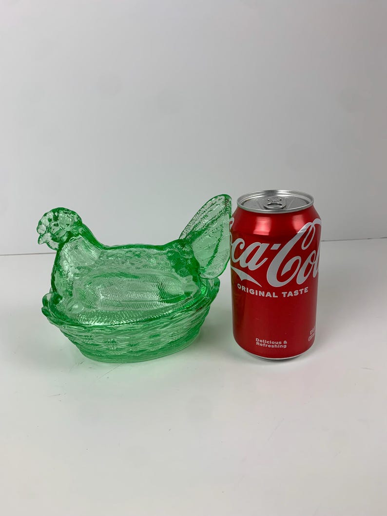 May include: A green glass hen-shaped dish with a textured surface. The dish is next to a red Coca-Cola can. The hen has a detailed head, wings, and tail, and sits atop a basket-like base.