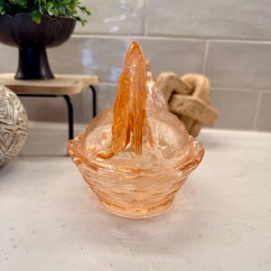 May include: A peach-colored, vintage-style glass candy dish with a handle. The dish has a textured, patterned design and a scalloped edge. The handle is a vertical, pointed shape. The dish is sitting on a white surface.