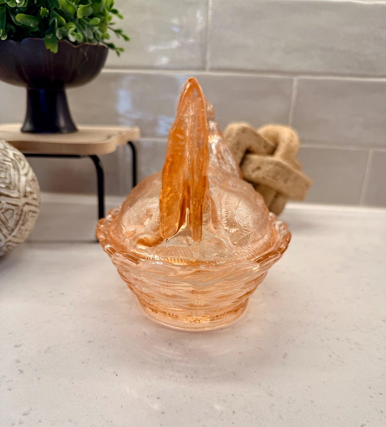 May include: A peach-colored, vintage-style glass candy dish with a handle. The dish has a textured, patterned design and a scalloped edge. The handle is a vertical, pointed shape. The dish is sitting on a white surface.