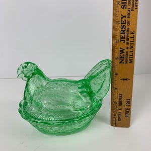 May include: A green glass hen-shaped dish with a textured surface. The dish is approximately 6 inches long. A wooden ruler with text is next to the dish. The text on the ruler says "NEW JERSEY SILICA".