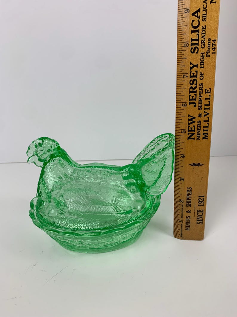 May include: A green glass hen-shaped dish with a textured surface. The dish is approximately 6 inches long. A wooden ruler with text is next to the dish. The text on the ruler says "NEW JERSEY SILICA".