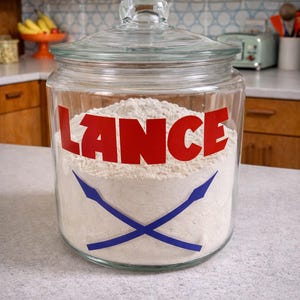 Pode incluir: Um frasco de vidro transparente cheio de farinha branca, com a etiqueta "LANCE" em letras vermelhas. Dois gráficos azuis de lanças cruzadas estão abaixo. O frasco tem uma tampa de vidro.