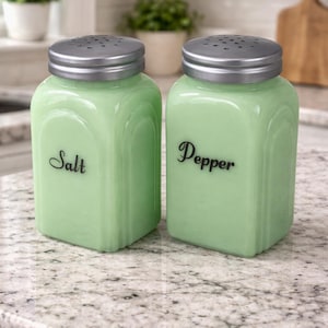 May include: A pair of vintage-style salt and pepper shakers. The square, light green glass jars have silver-colored metal lids with small holes. The word "Salt" is written on one shaker, and "Pepper" on the other in a black script font.