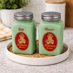 Op de afbeelding: Twee lichtgroene glazen zout- en peperstrooiers met zilveren deksels. Elke strooier heeft een rood-wit etiket met een vintage illustratie en de tekst "Sunbeam Bread". De strooiers staan op een wit dienblad.