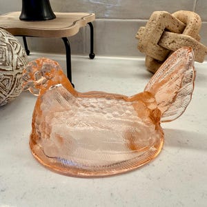 May include: A decorative, light pink glass hen-shaped dish. The dish has a textured surface and is designed to resemble a chicken. The hen has a detailed wing and tail. The dish is likely used for serving or display.