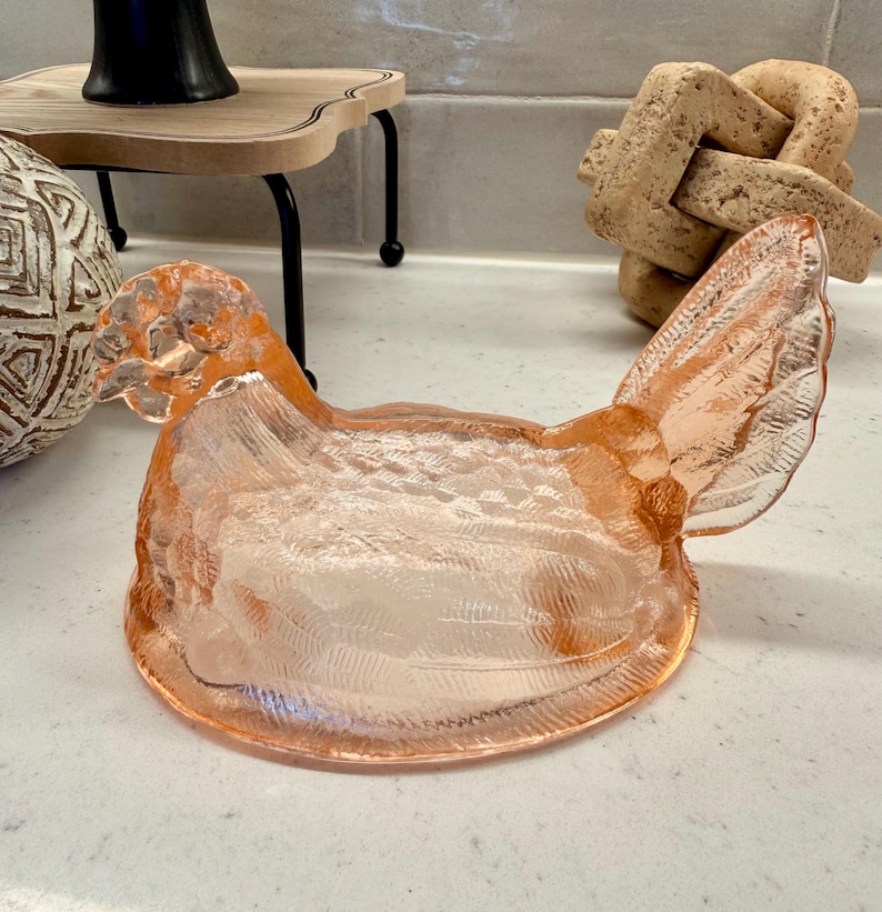 May include: A decorative, light pink glass hen-shaped dish. The dish has a textured surface and is designed to resemble a chicken. The hen has a detailed wing and tail. The dish is likely used for serving or display.