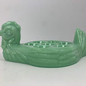 Pode incluir: Um porta-ovos de vidro verde em forma de galinha, com seis reentrâncias para os ovos.