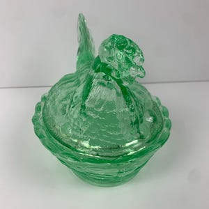 May include: A green glass hen-shaped covered dish. The dish features a textured surface and a detailed hen design, including a head, tail, and body. The lid fits snugly over the base, creating a decorative and functional piece.