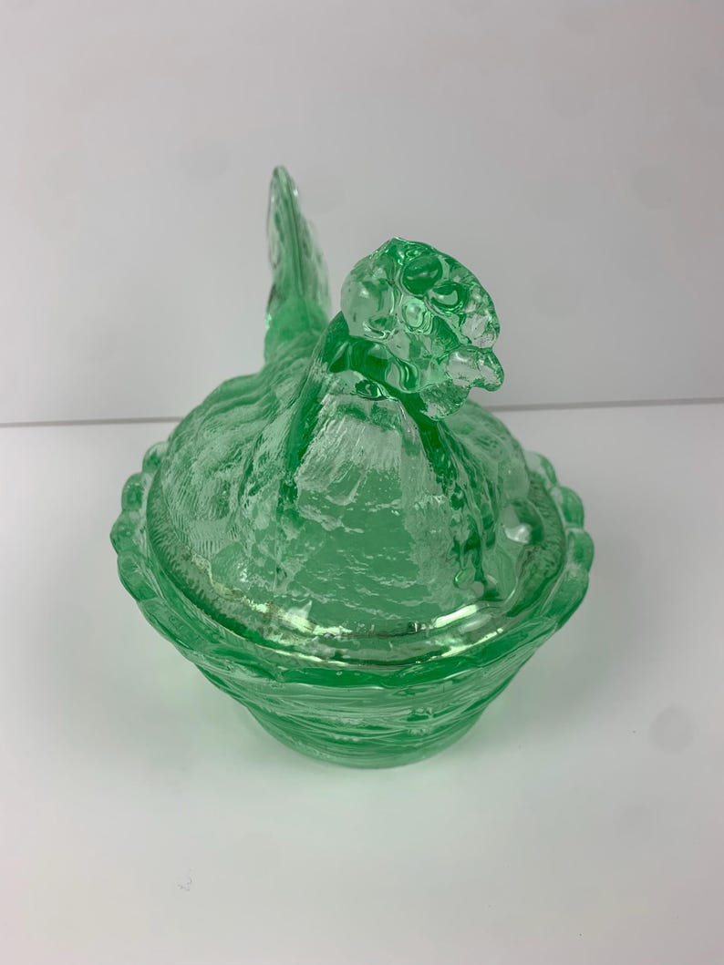 May include: A green glass hen-shaped covered dish. The dish features a textured surface and a detailed hen design, including a head, tail, and body. The lid fits snugly over the base, creating a decorative and functional piece.