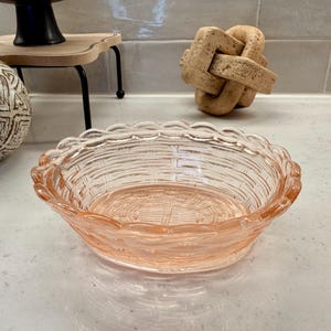 May include: A clear, pink glass bowl with a woven basket design. The bowl has a scalloped edge and is likely used for serving or display. The bowl is sitting on a white surface.