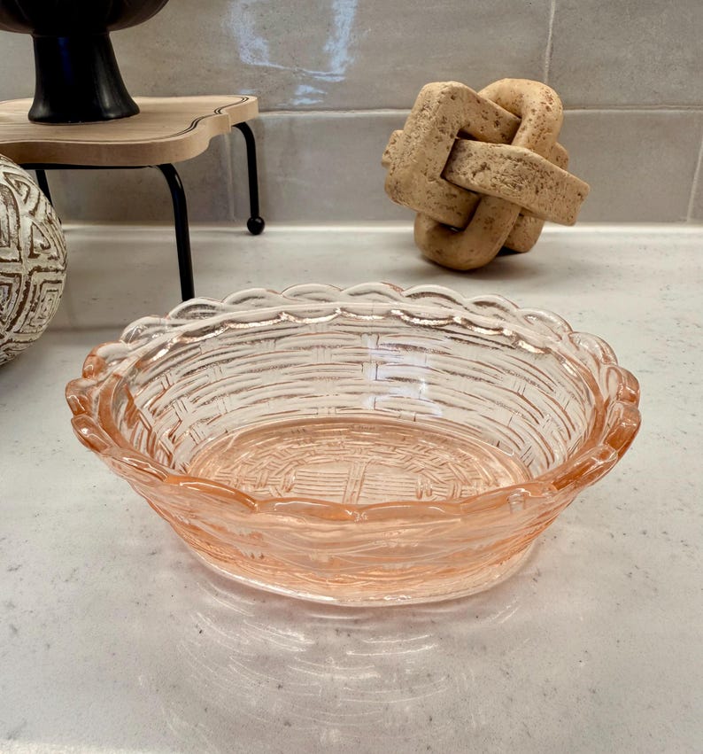 May include: A clear, pink glass bowl with a woven basket design. The bowl has a scalloped edge and is likely used for serving or display. The bowl is sitting on a white surface.