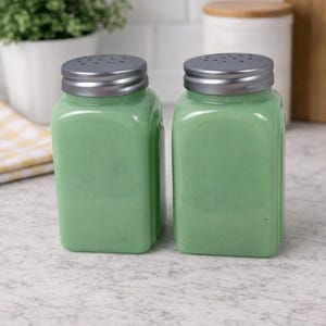 May include: Two green glass salt and pepper shakers with silver-colored lids. The shakers are square-shaped with rounded edges and sit on a light-colored countertop. A small plant in a white pot is in the background.