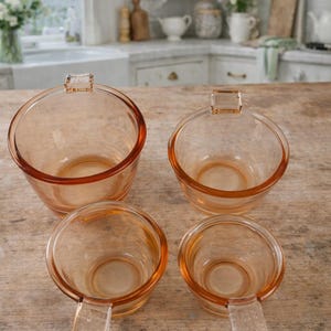 Può includere: Quattro ciotole vintage in vetro color pesca con manici. Le ciotole sono disposte su una superficie di legno, con uno sfondo di cucina visibile. Le ciotole hanno una forma arrotondata con un manico su un lato.