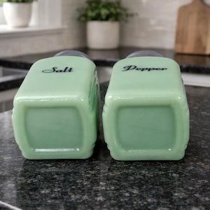May include: Two vintage, light green salt and pepper shakers. The rectangular shakers have rounded corners and black text that reads "Salt" and "Pepper". The shakers are on a dark speckled countertop.