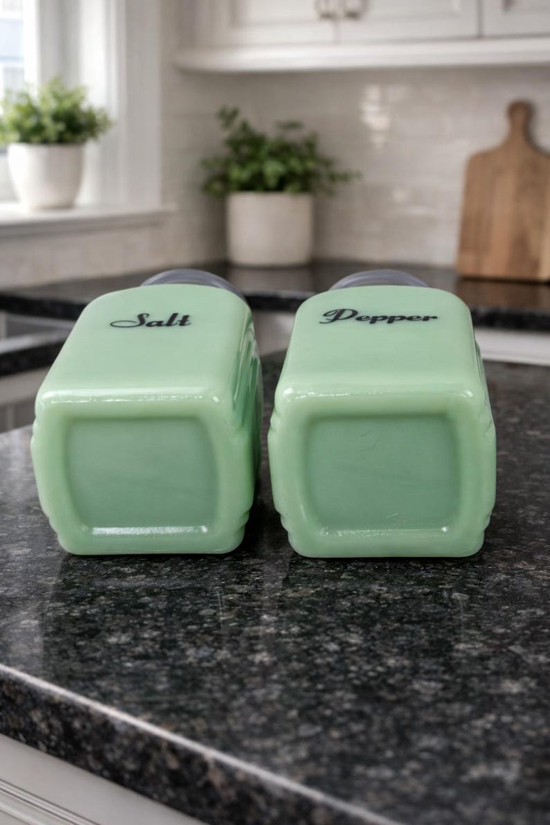 May include: Two vintage, light green salt and pepper shakers. The rectangular shakers have rounded corners and black text that reads "Salt" and "Pepper". The shakers are on a dark speckled countertop.