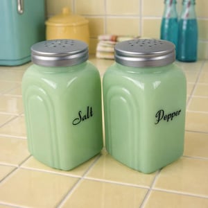 May include: A pair of vintage-style, light green glass salt and pepper shakers with silver lids. The word "Salt" is written on one shaker, and "Pepper" on the other. The shakers are on a yellow tiled surface.