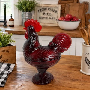Può includere: Un piatto con coperchio in vetro rosso scuro a forma di gallo, caratterizzato da un motivo a piume testurizzato sul corpo e una cresta rosso brillante. L'oggetto poggia su una base circolare e include un coperchio rimovibile superiore.