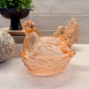 May include: A peach-colored glass hen-shaped dish with a textured surface. The hen is sitting in a basket-like base. The dish is likely used for decorative purposes or to hold small items.