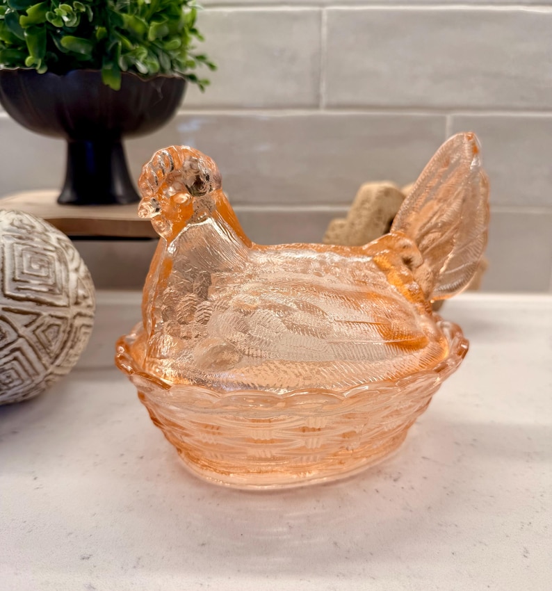May include: A peach-colored glass hen-shaped dish with a textured surface. The hen is sitting in a basket-like base. The dish is likely used for decorative purposes or to hold small items.