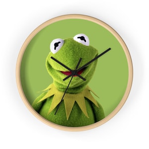May include: A round wall clock with a light wooden frame featuring a green background and a central image of a green felt frog character with large white eyes, a wide smile, and a pointed collar. The clock has three thin black hands for timekeeping.