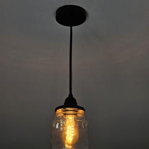 Könnte beinhalten: Eine Hängeleuchte mit einem Lampenschirm in Form eines transparenten Glasbehälters, schwarzen Metallbeschlägen und einer warm getönten Glühbirne. Die Leuchte hängt an einem schwarzen Kabel und einer Deckenhalterung.