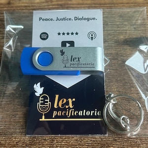 May include: A blue and silver USB flash drive, branded with "lex pacificatoria", is packaged with a business card and a keychain. The card features a dove and microphone graphic, along with the same text. The packaging is clear plastic.