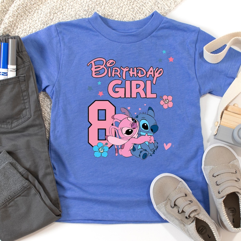 以下が含まれることがあります： ピンク色の「Birthday Girl」の文字と数字の8が入った青いTシャツ。Tシャツには、花と星のディテールとともに、漫画のキャラクター、スティッチとエンジェルが抱き合っている様子が描かれています。グレーのズボンと靴も写っています。