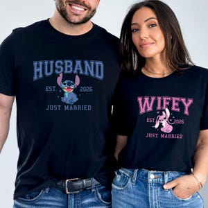 Op de afbeelding: Twee bijpassende zwarte t-shirts. E&eacute;n t-shirt met het woord "HUSBAND" in blauw met een cartoonfiguur, de andere met "WIFEY" in roze. Beide hebben de tekst "EST. 2026 JUST MARRIED".