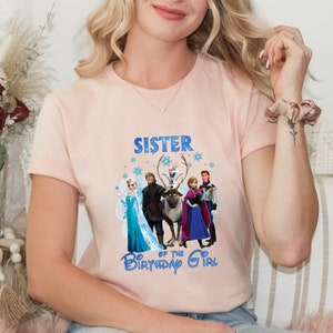 May include: A pale pink t-shirt with a Frozen-themed graphic. The design includes characters from the film and the text "SISTER" and "OF THE BIRTHDAY GIRL" in blue. The shirt has short sleeves.