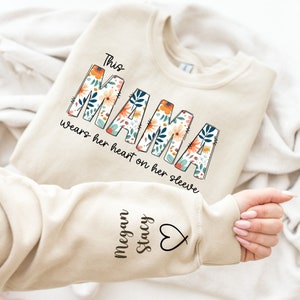 May include: Beige sweatshirt with the text "This MAMA wears her heart on her sleeve" in a floral design. The sleeve features the names "Megan Stacy" and a heart symbol. The sweatshirt is folded on a white surface.
