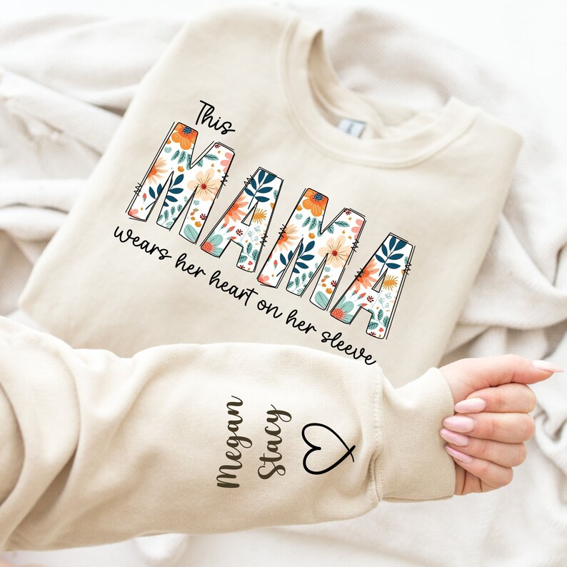 May include: Beige sweatshirt with the text "This MAMA wears her heart on her sleeve" in a floral design. The sleeve features the names "Megan Stacy" and a heart symbol. The sweatshirt is folded on a white surface.