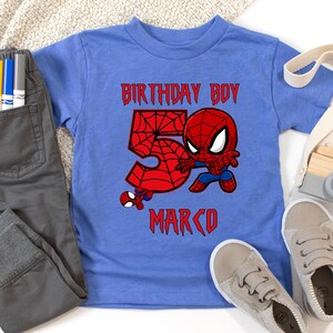 May include: A blue t-shirt with a Spider-Man graphic and the text "BIRTHDAY BOY 5 MARCO" in red. The image includes grey trousers and light grey trainers. The design features a large red number 5.