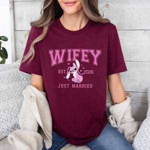Op de afbeelding: Bordeauxrood T-shirt met het woord "Wifey" in roze blokletters, het jaar "2026" en de woorden "Just Married". Een roze stripfiguur is afgebeeld onder de tekst. Het shirt heeft korte mouwen.