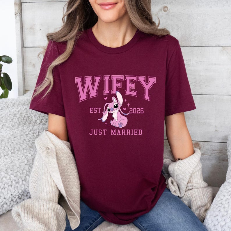 Op de afbeelding: Bordeauxrood T-shirt met het woord "Wifey" in roze blokletters, het jaar "2026" en de woorden "Just Married". Een roze stripfiguur is afgebeeld onder de tekst. Het shirt heeft korte mouwen.