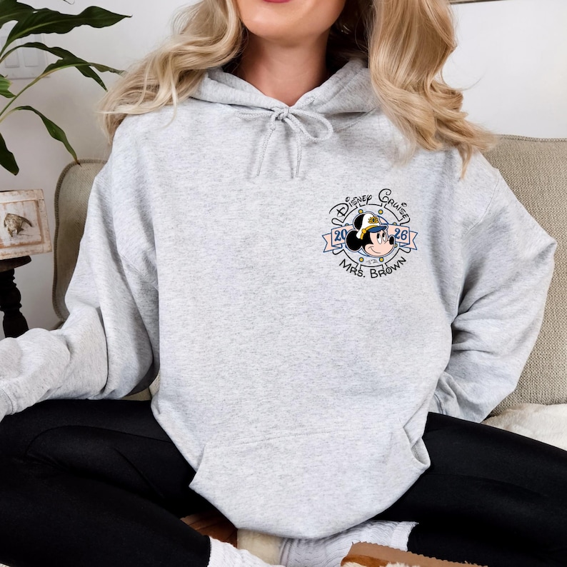 Op de afbeelding: Een lichtgrijze hoodie met een trekkoord. De hoodie heeft een cirkelvormige afbeelding met Mickey Mouse met een matrozenhoed, de woorden "Disney Cruise 2026" en "Mrs. Brown".