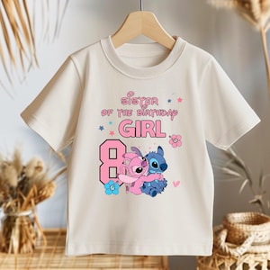 以下が含まれることがあります： ピンク色の「Sister of the Birthday Girl」の文字と大きなピンク色の数字8がプリントされた、クリーム色のTシャツ。青とピンクのアクセントで、2人の漫画のキャラクターが抱き合っているグラフィックが描かれています。木製のハンガーにかかっています。