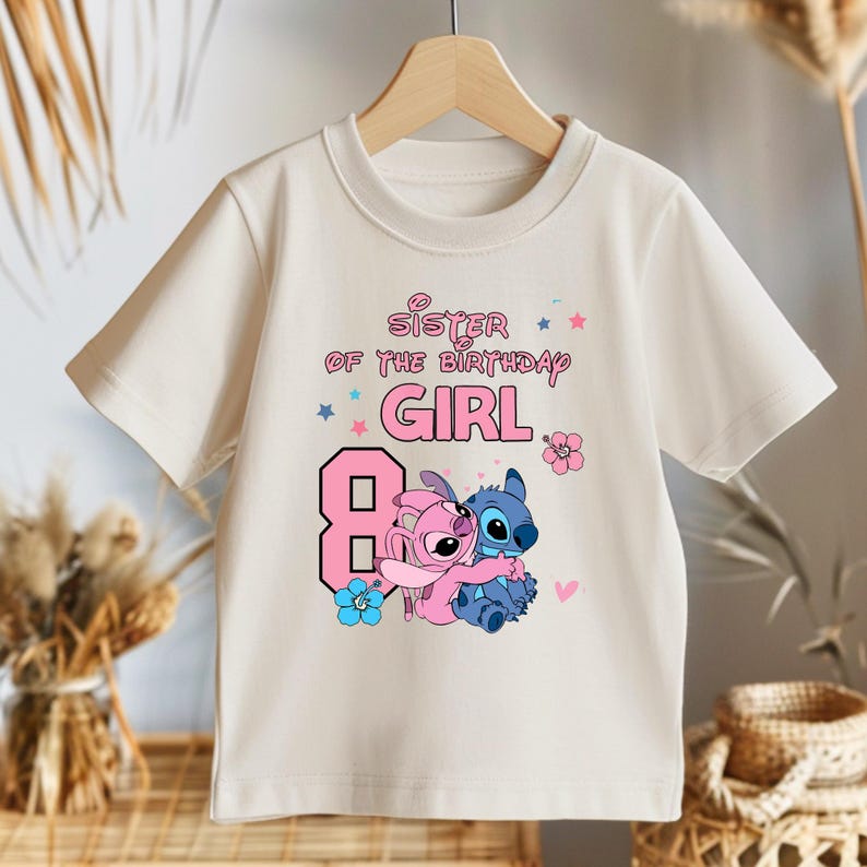 以下が含まれることがあります： ピンク色の「Sister of the Birthday Girl」の文字と大きなピンク色の数字8がプリントされた、クリーム色のTシャツ。青とピンクのアクセントで、2人の漫画のキャラクターが抱き合っているグラフィックが描かれています。木製のハンガーにかかっています。